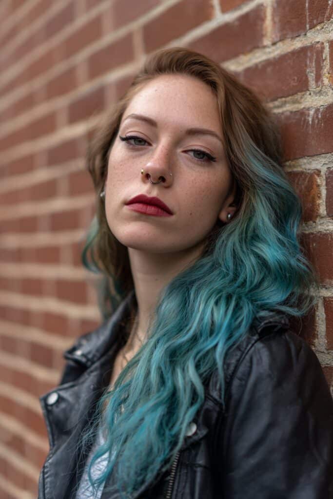 American woman with long wavy mermaid hair in teal and purple tones, glam rocker makeup against urban brick wall.