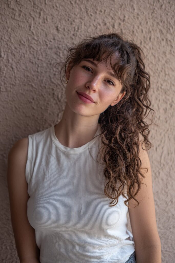 Casual portrait of American model with curly ponytail, face-framing curtain bangs against textured plaster wall backdrop.