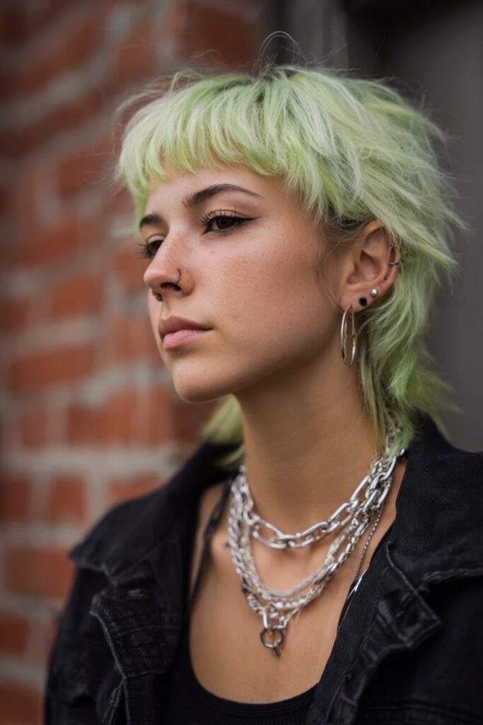 American model with neon green spiky emo shag haircut, side-swept fringe, heavy eyeliner, chain accessories, brick wall background.