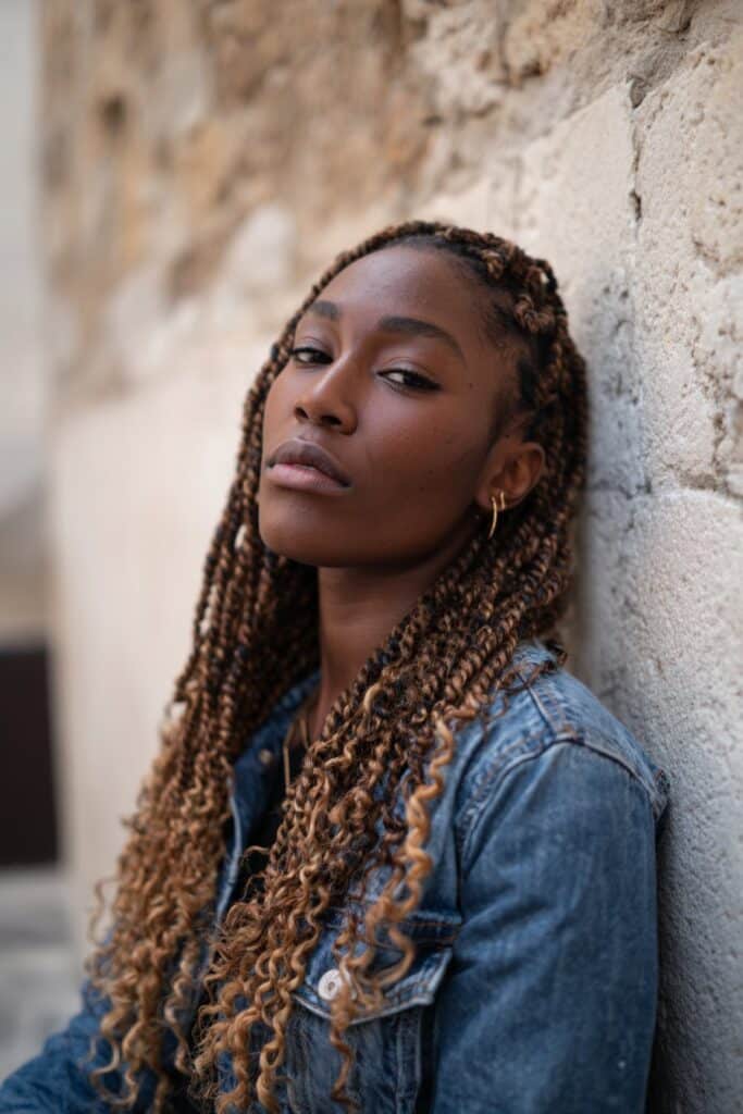 Fashionable Black woman with long ombré crochet faux locs, neutral makeup, denim jacket, textured backdrop.