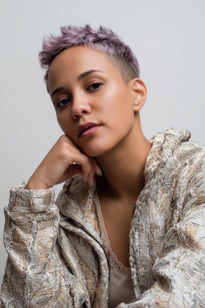 Fashion-forward American model with pastel purple spiky pixie haircut wearing textured jacket against seamless white studio background.