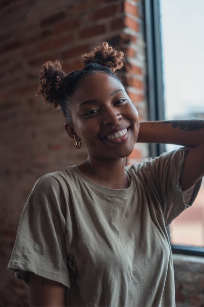 Playful portrait of Black American woman with short 4c hair in two curly pigtails against urban brick wall.