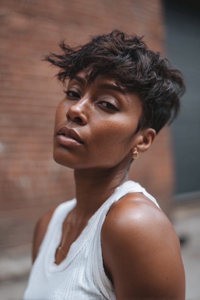 Textured portrait of Black woman with choppy layered pixie cut, tousled movement, natural makeup against brick wall.