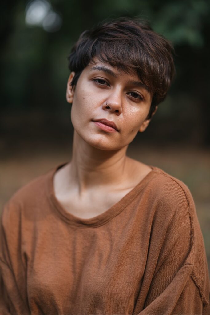 American woman with razored messy pixie layers, highlighted ends, natural glam look, Instagram-style beauty against gray fabric.