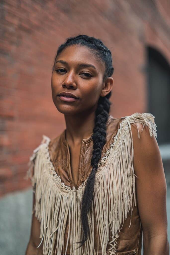 American woman with textured pull-through braid, festival outfit with fringe details, glowing skin against brick wall.