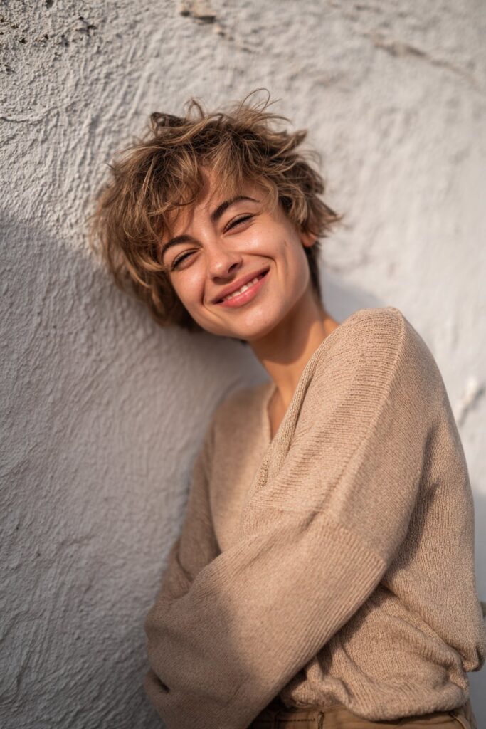 Natural portrait of American woman with voluminous shaggy stacked pixie, tousled layers against textured plaster wall.