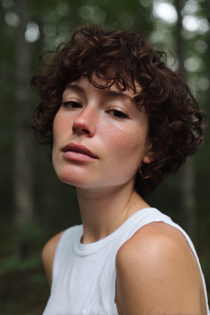 American woman with messy pixie and soft curtain bangs, natural dark blonde tone, light sweater in urban forest.