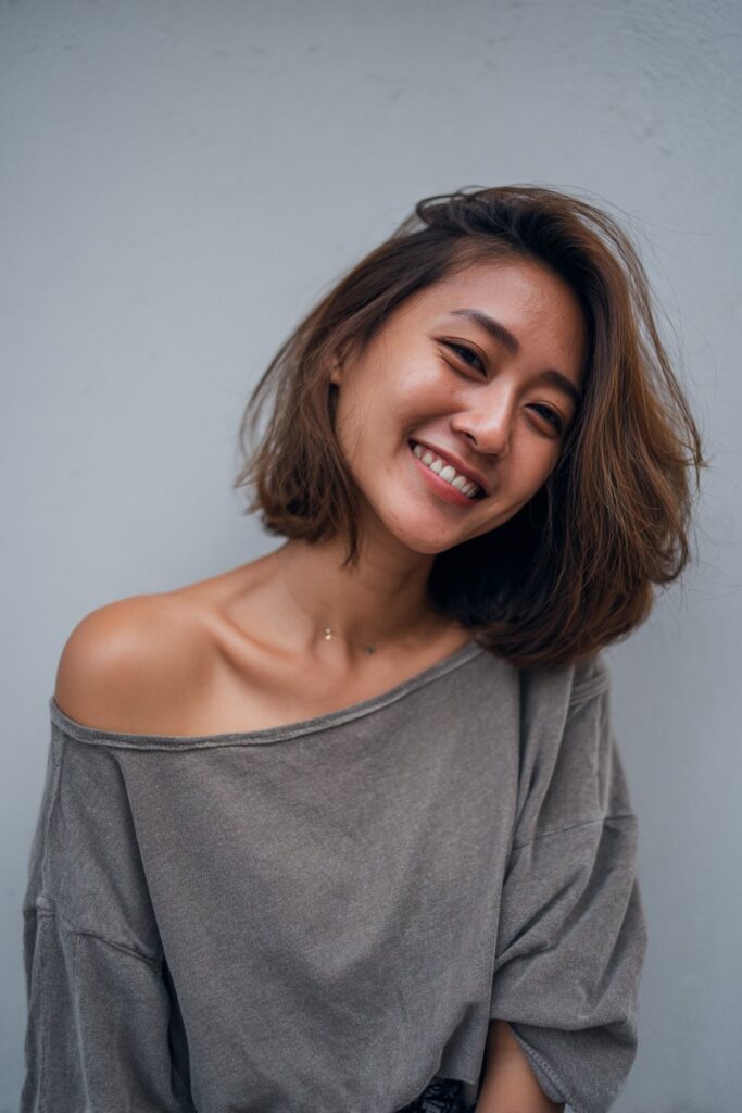 American woman with butterfly haircut featuring balayage highlights, friendly expression, trendy clothing against soft gray fabric backdrop.