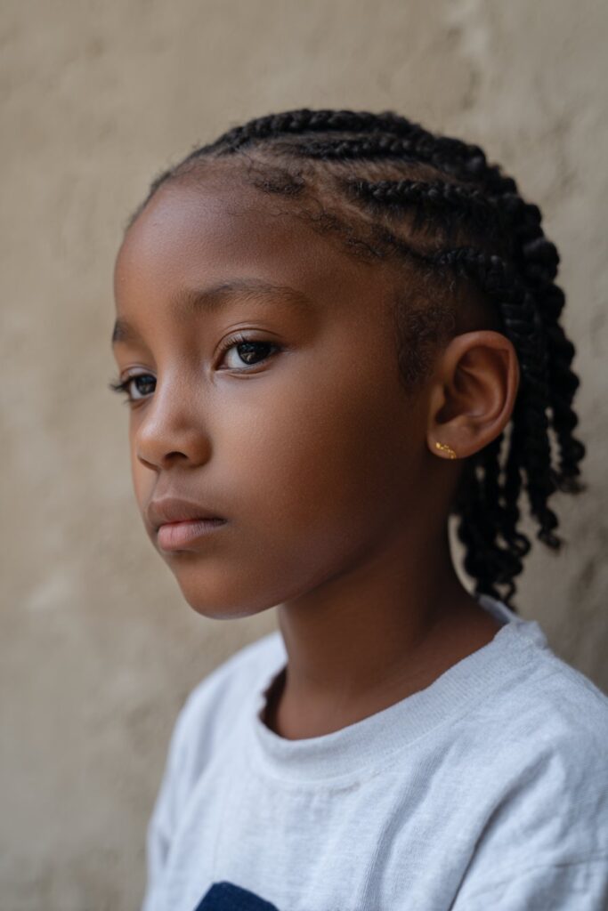 American kid with neatly braided side-parted cornrows in simple elegant pattern, minimal accessories, photographed against textured plaster backdrop.