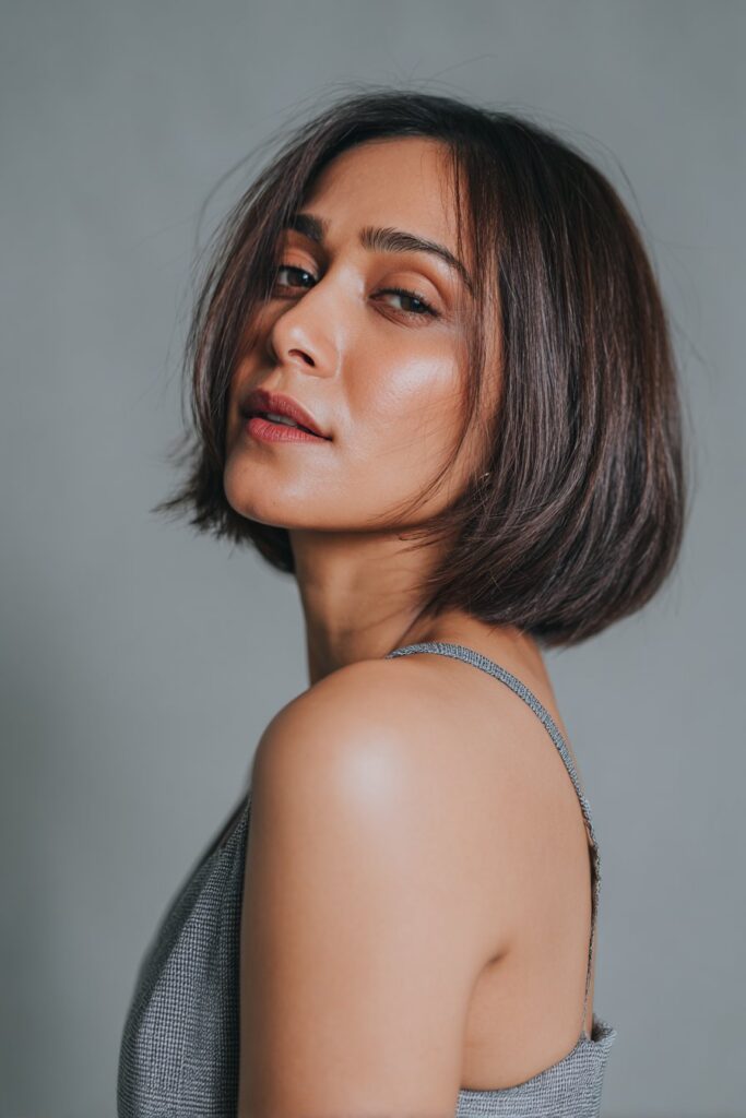 American woman with sleek butterfly haircut, sharp layers, polished finish, modern outfit, confident gaze, gray backdrop.