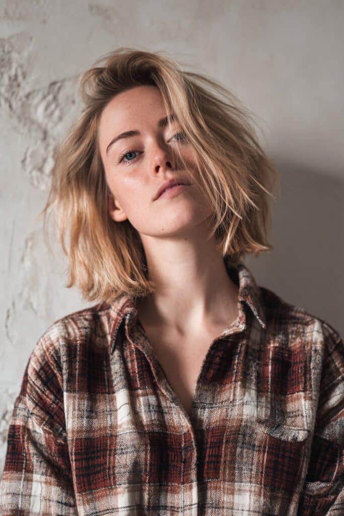 American woman with soft tousled blonde hair, 90s grunge style oversized flannel shirt against textured plaster wall backdrop.