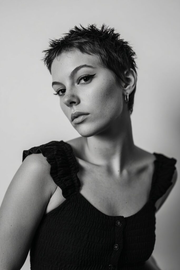 American female model with spiky short crop textured top, bold eyeliner, edgy attitude, simple monochrome outfit, white backdrop.