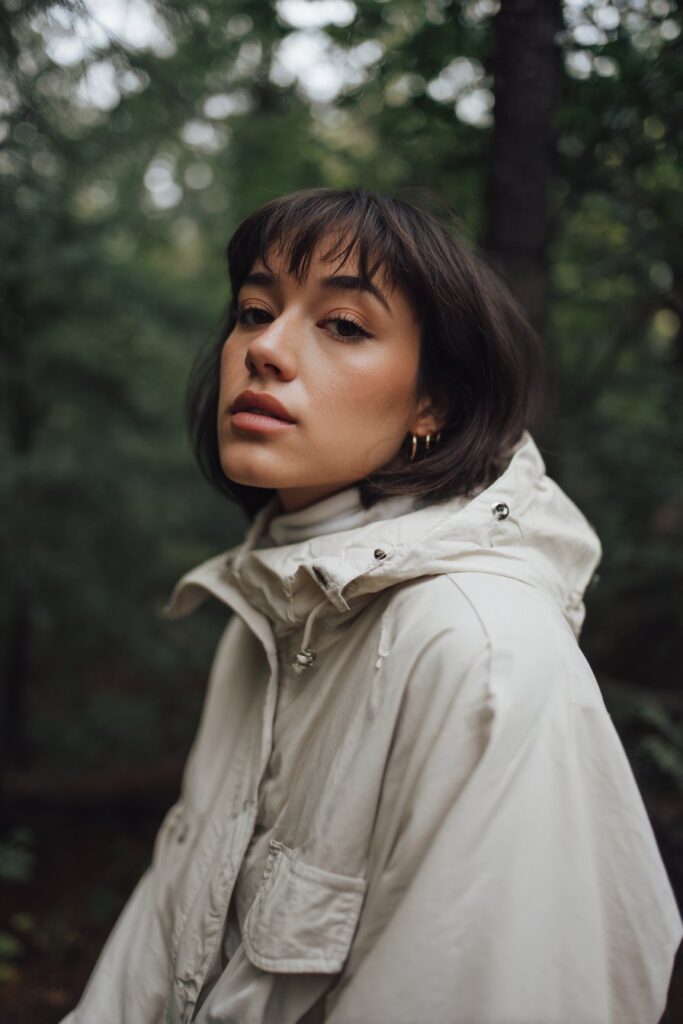 Polished portrait of American model with stacked pixie featuring curtain bangs, neutral makeup in forest setting.