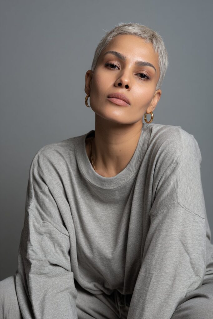 Modern portrait of American model with sleek stacked pixie, long sideburns, monochrome outfit against gray fabric.
