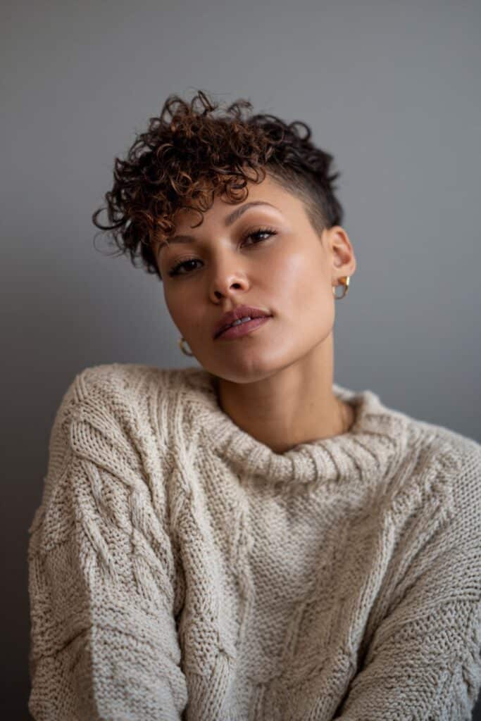 Cozy portrait of American model with naturally curly stacked pixie, warm makeup, knitwear against gray fabric.