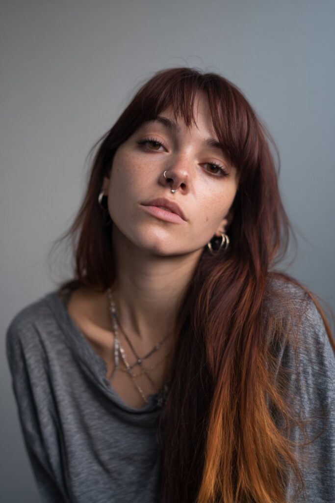 American female model with sunset ombre shag in red-orange-yellow shades, thick bangs, flicked ends, emo accessories, gray backdrop.