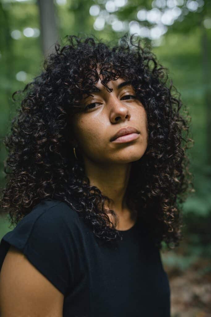 Bold portrait of American model with ultra-voluminous curls, dramatic curtain bangs, neutral outfit in natural forest setting.