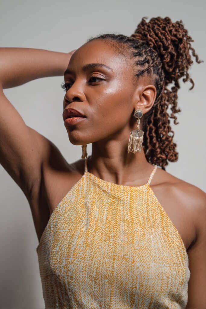 Black woman with crochet curls in high ponytail, sleek edges, bold earrings, white seamless backdrop.