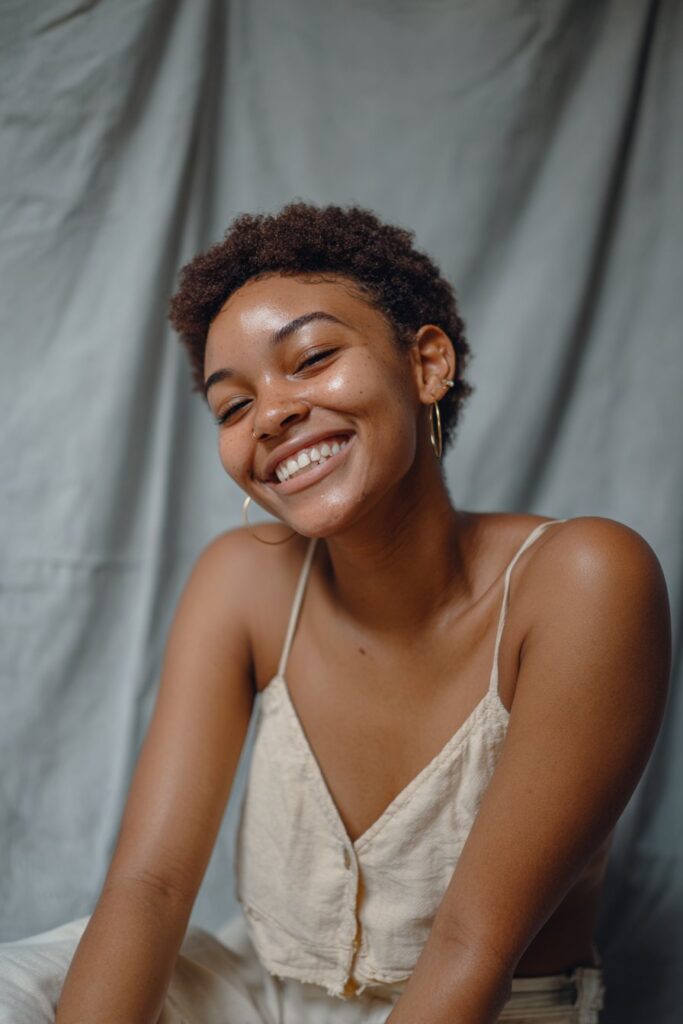 Black woman with cropped cut styled in perm rod curls, bouncy texture, joyful smile, casual fashion, gray backdrop.