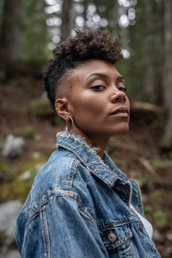 Black woman with bold high top fade, clean sculpted lines, vintage denim jacket, natural forest backdrop, outdoor lighting.