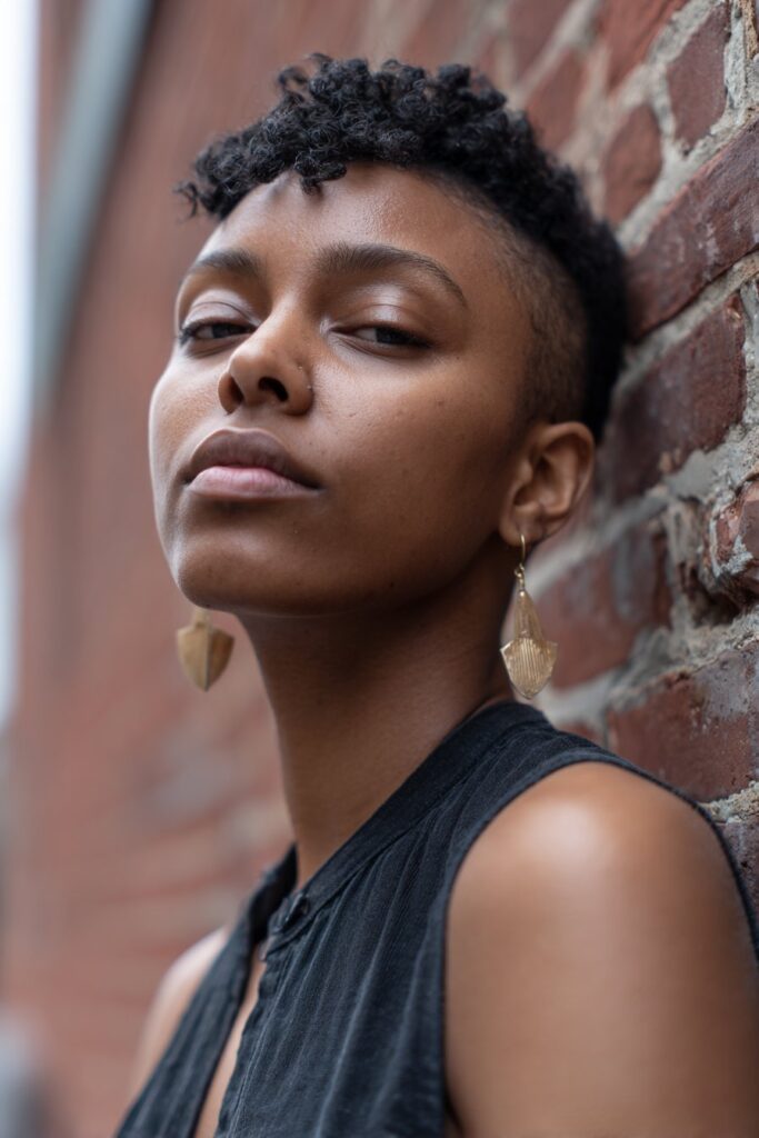 Stylish Black woman with tapered natural fade, tight curls on top, clean faded sides, minimalist earrings, urban brick wall.