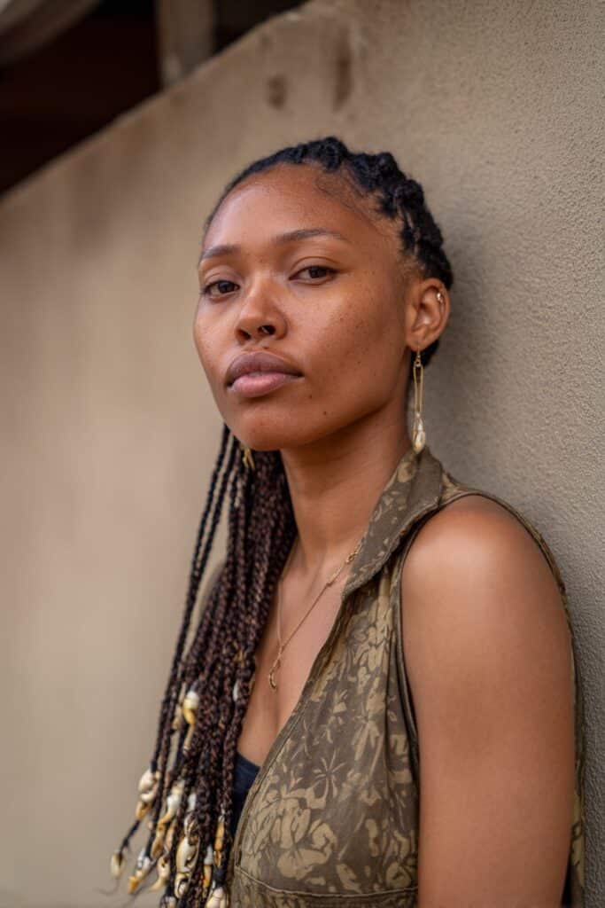 American woman with tribal braids decorated with cowrie shells, radiant skin tone, earthy-toned outfit, subtle gold highlighter backdrop.