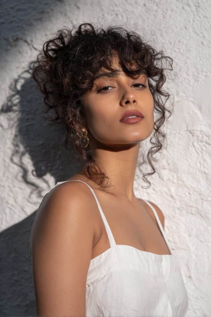 Elegant portrait of American woman with curly updo, cascading curtain bangs, warm makeup against textured plaster wall.