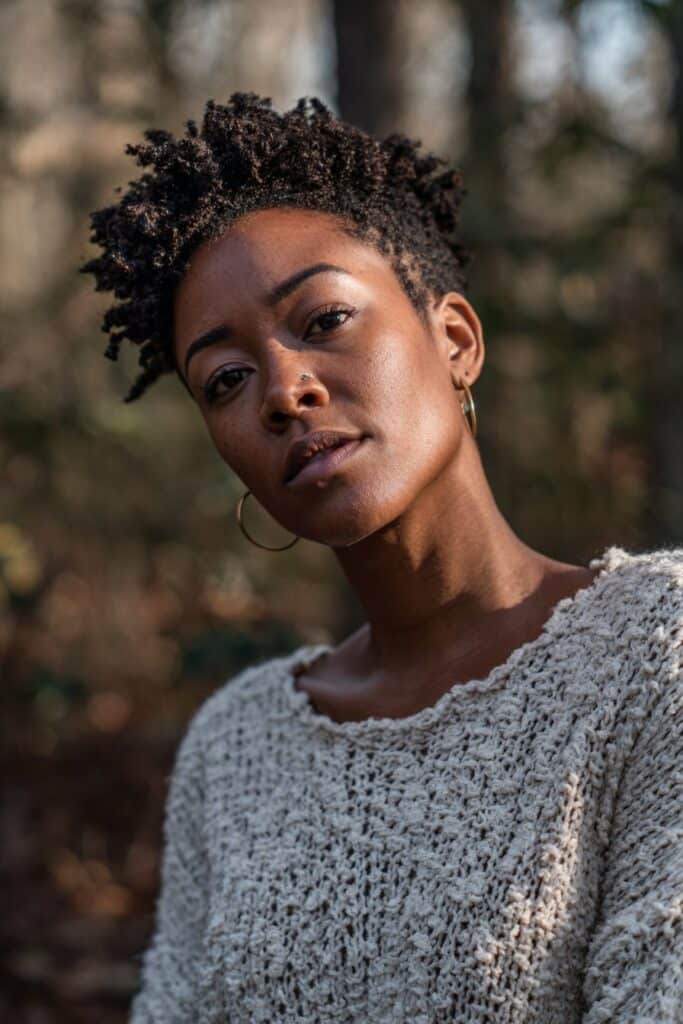 American female model with 4C hair pixie cut, soft waves and coils, radiant skin, textured knit top, forest backdrop.