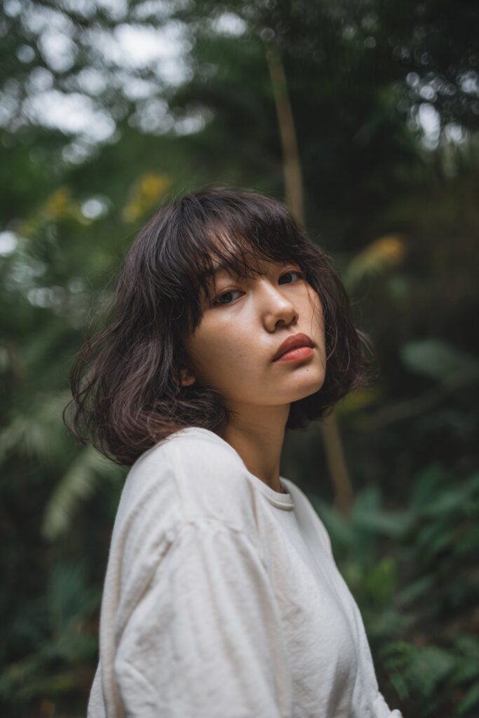 American woman with chin-length shaggy bob and curtain fringe bangs, fresh natural look, soft outfit in forest scene.