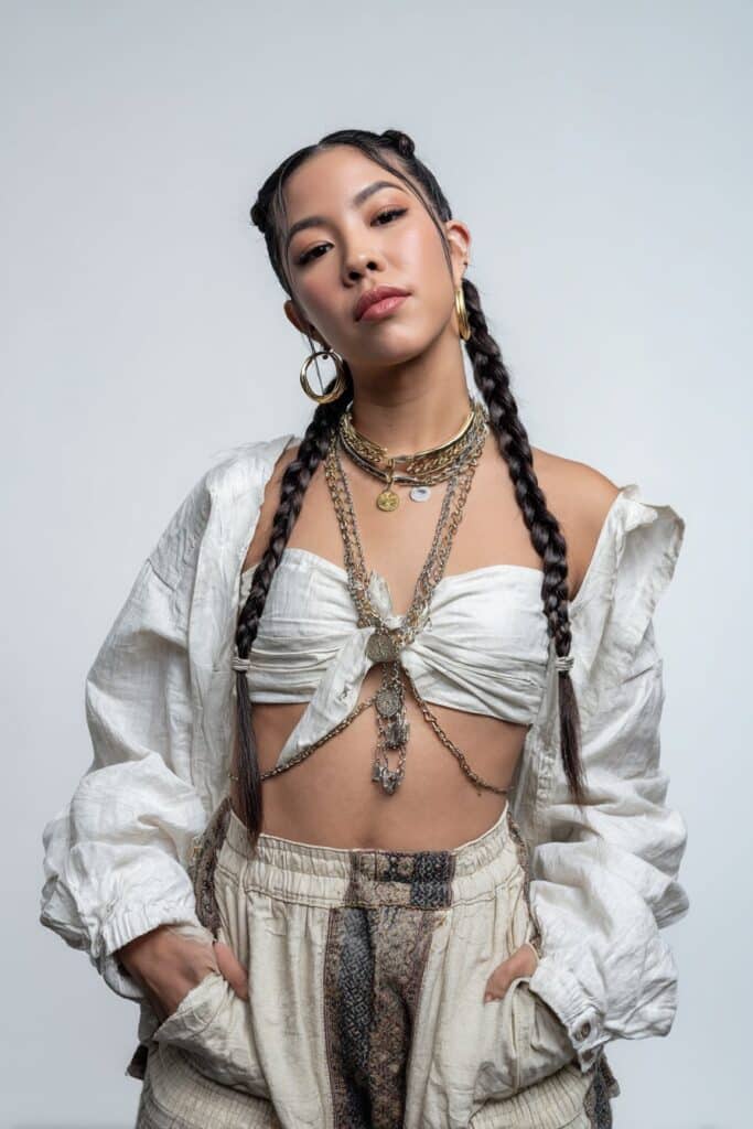 Portrait of American woman with milkmaid braids, confident expression, concert outfit, layered accessories on white backdrop.