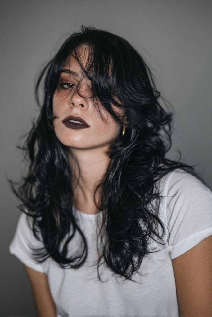 American model with long layered hair parted deeply to one side, fringe sweeping over one eye, soft waves and black hair with subtle shine, moody gray fabric backdrop.