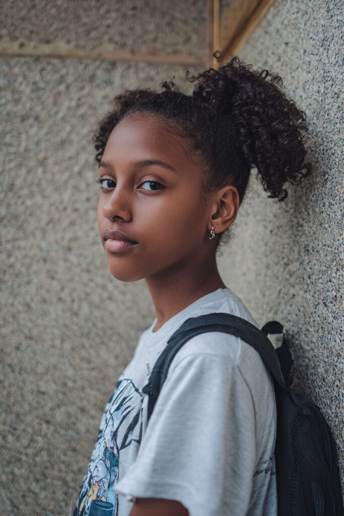 Confident teen girl with curly faux hawk hairstyle, pinned sides and curly volume on top, posing against a classroom wall.