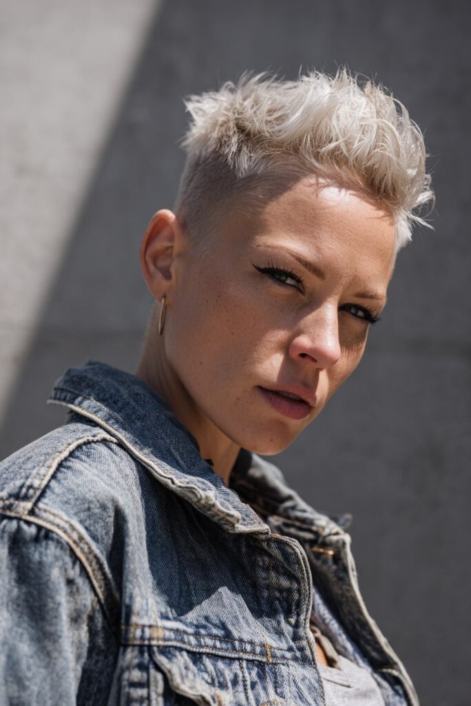 Edgy faux hawk-style pixie cut with spiked top and closely shaved sides, icy blonde hair, dramatic eyeliner, worn with a denim jacket on a soft gray backdrop.
