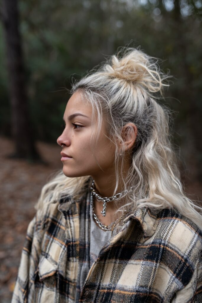 Grunge-inspired hairstyle featuring a messy half-up top knot on bleached hair with visible dark roots, loose bottom strands and flannel shirt, shot outdoors at forest edge.