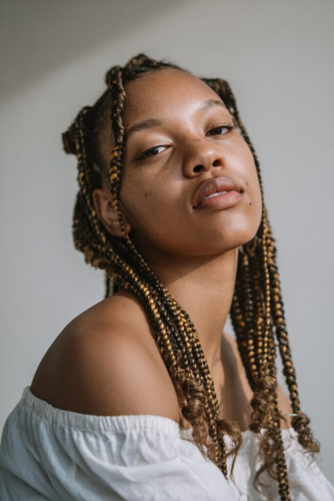 Jayda Wayda-inspired look with two thick back braids ending in spiral curls, worn by a Black woman in a casual tank top at the edge of a forest.