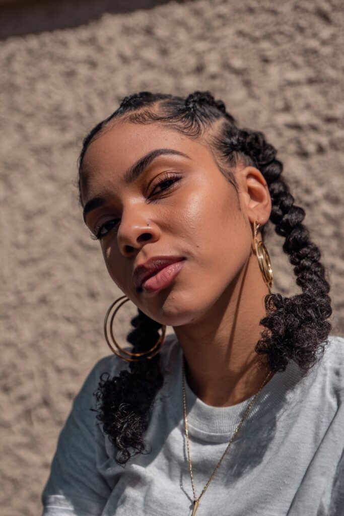 Short braided bob with curly ends in a Jayda Wayda-inspired look, worn by a Black woman with a defined jawline and hoop earrings, soft plaster wall behind.