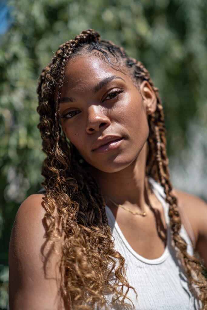 African-American model with Jayda Wayda-inspired boho braids blended with curly texture throughout, golden brown accents, styled in an outdoor forest scene.
