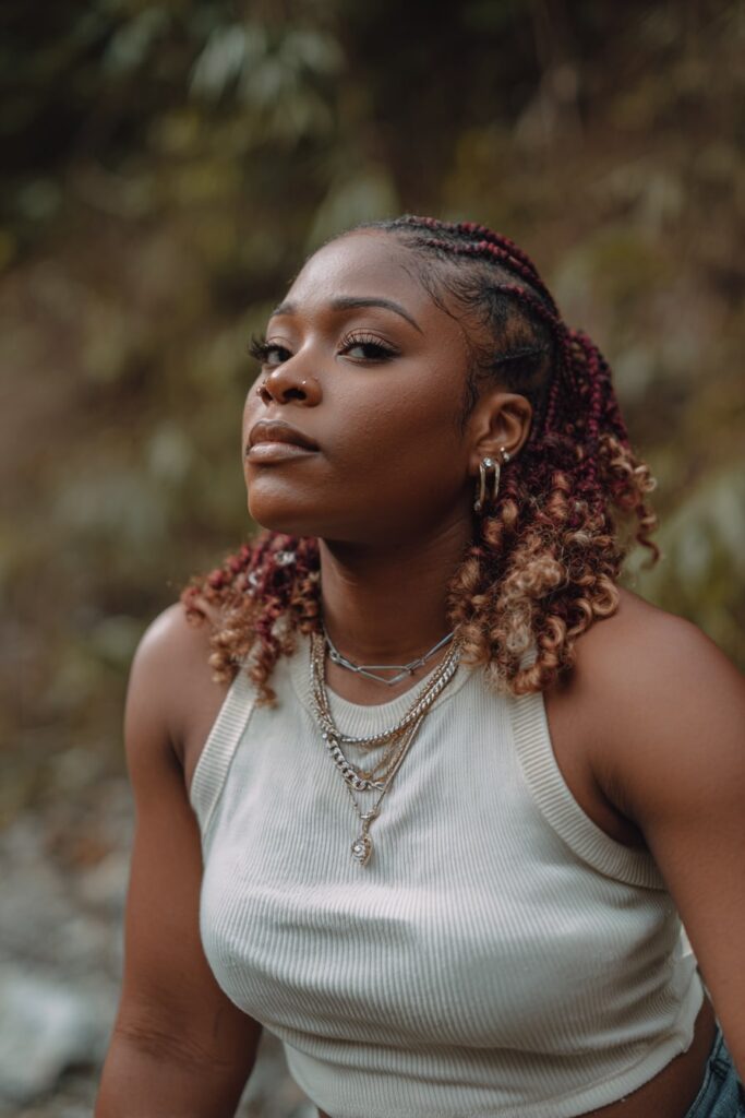 Jayda Wayda braid look with curly ends and burgundy + blonde color accents, styled with soft glam makeup and a natural forest edge setting.