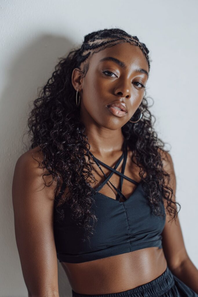 Black model with crisscross cornrow pattern and long curly braids in Jayda Wayda style, baby hairs styled cleanly, neutral-toned lips, on a white background.