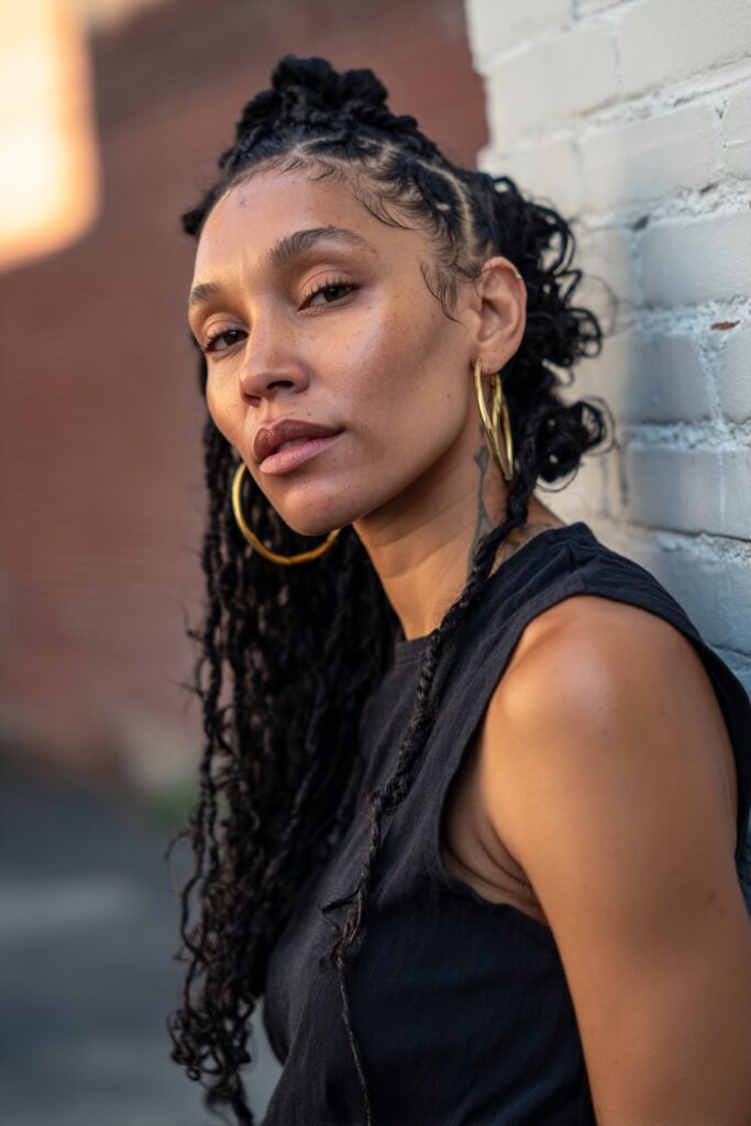 Black American model with long Jayda Wayda braids and heart-shaped cornrows on each side, finished with curly ends, laid baby hairs, and hoop earrings in front of a brick wall.