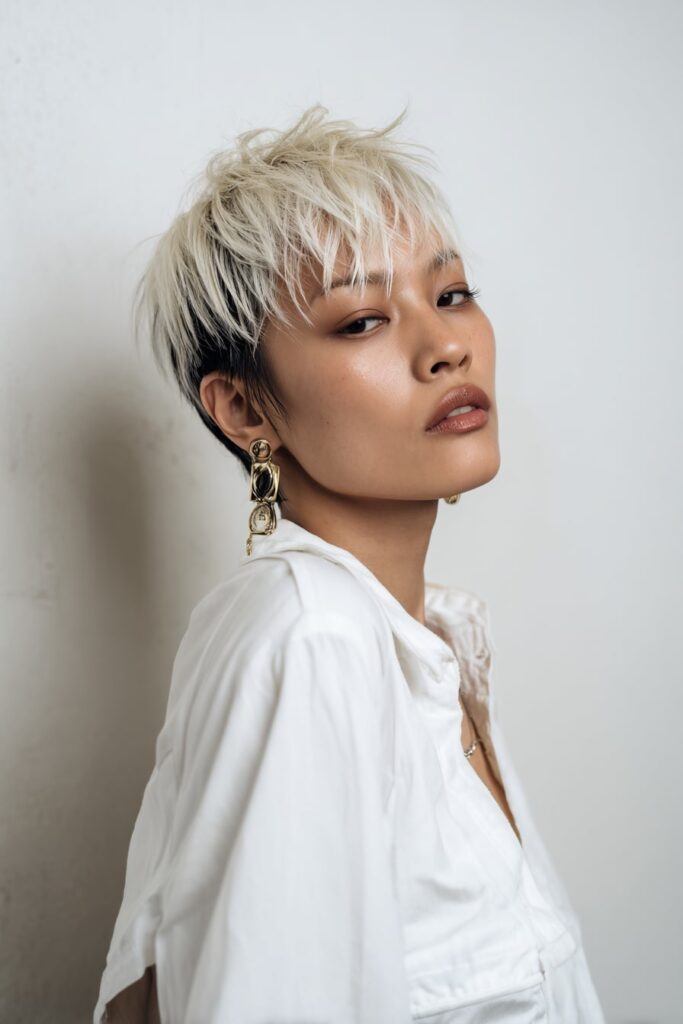 Bold two-toned pixie haircut with a platinum top and dark undercut, choppy fringe and short sides, styled with neutral makeup against a white studio backdrop.