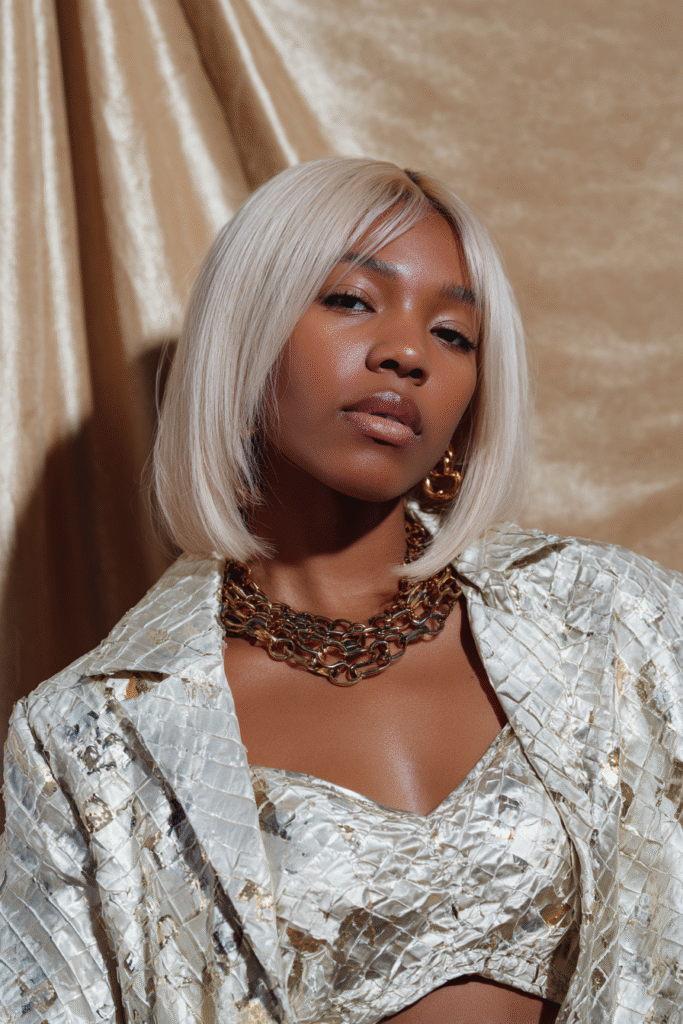 A vibrant female model in her 20s with dark skin and shoulder-to-waist-length straight hair in 613 blonde, styled in a sleek baddie wig with a glossy finish, framed from chest-level upward, photographed against a seamless soft gold backdrop with a satin finish. She has bold makeup, a trendy outfit, a warm skin tone, and a confident expression, captured with soft natural lighting.