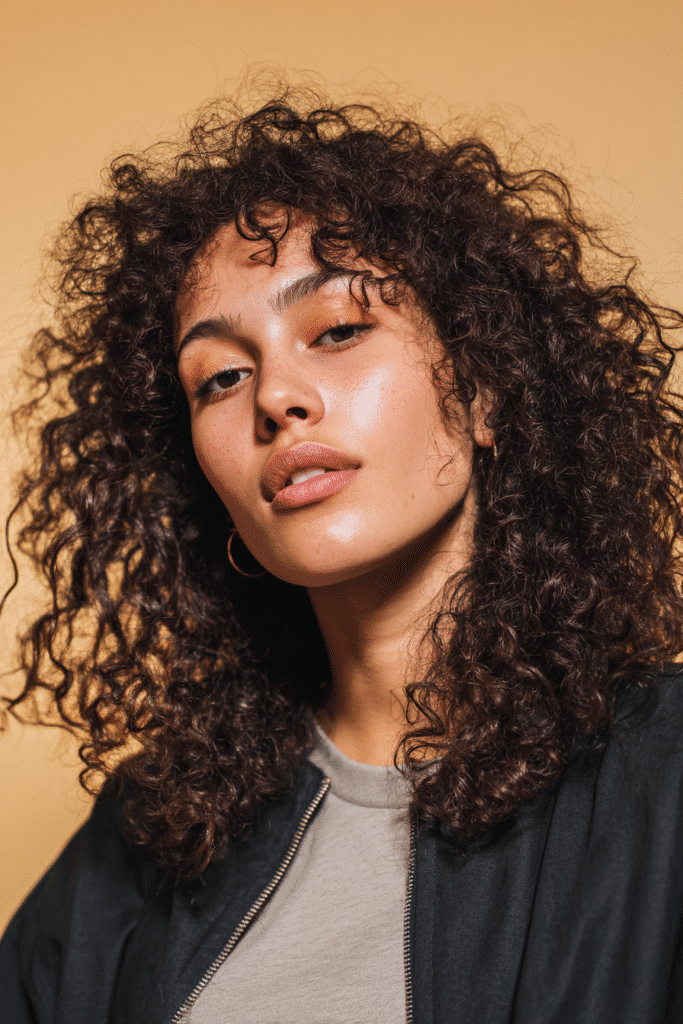 A vibrant female model in her 20s with shoulder-to-waist-length curly half wig, styled with an afro curly volume style and full, bouncy curls flowing down, photographed against a seamless light amber backdrop with a satin finish. She has subtle makeup, a trendy outfit, a warm skin tone, and a friendly expression, captured with soft natural lighting.