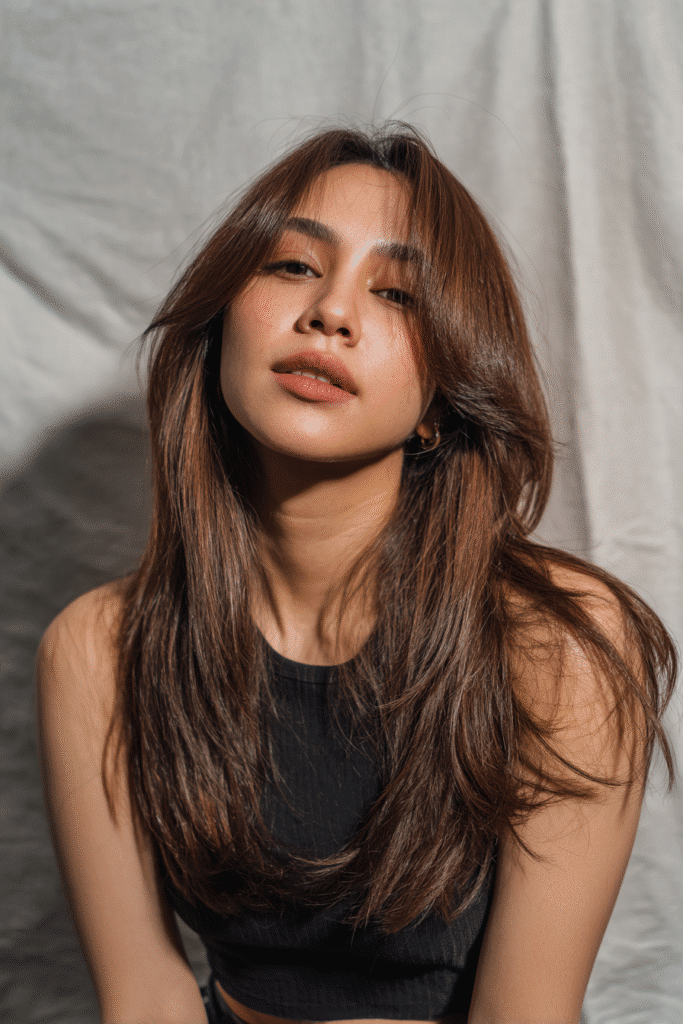 A vibrant American female model in her 20s with chest-length asymmetrical Hime Cut long hair featuring uneven layers, bold sidelocks, and side bangs, styled dynamically, photographed against a seamless light charcoal backdrop with a satin texture. She has subtle makeup, a trendy outfit, a warm skin tone, and a friendly expression, captured with soft natural lighting.