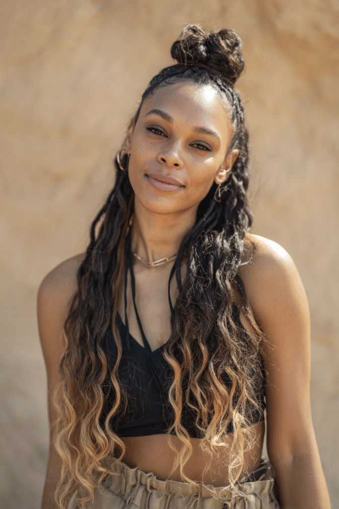 A vibrant Black female model in her 20s with shoulder-to-waist-length balayage wig with clip-ins, styled in a half-up twisted bun with balayage strands in caramel to honey blonde flowing down, photographed against a seamless warm sand backdrop with a matte texture. She wears subtle makeup, a trendy outfit, and has a warm skin tone with a friendly expression, captured with soft natural lighting.