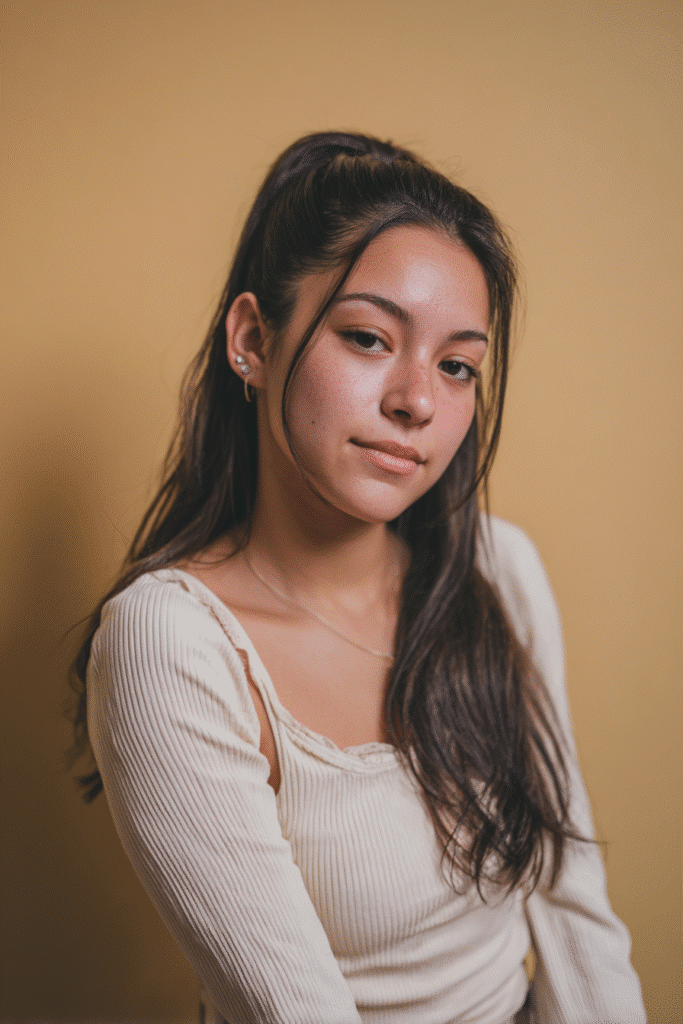 A vibrant white female model in her 20s with shoulder-to-waist-length straight wig, styled in a half-up high Barbie ponytail with quick weave strands flowing down, photographed against a seamless warm beige backdrop with a glossy sheen. She wears subtle makeup, a trendy outfit, and has a warm skin tone with a friendly expression, captured with soft natural lighting.