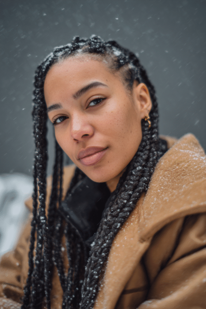 A vibrant female model in her 20s with shoulder-to-waist-length box braids, styled with neatly parted sections and a bold protective finish, captured in a close-up shot focusing on the hair, photographed against a seamless soft charcoal backdrop with a velvet texture. She has subtle makeup, a stylish winter coat, a warm skin tone, and a confident expression, captured with cozy winter lighting and soft snowflakes.