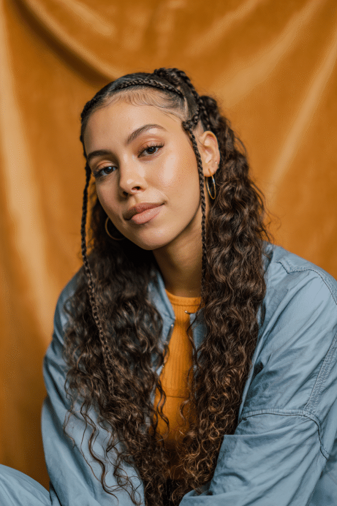A vibrant female model in her 20s with shoulder-to-waist-length wavy half wig, styled with intricate braids at the front and loose waves flowing down, photographed against a seamless warm amber backdrop with a glossy sheen. She wears subtle makeup, a trendy outfit, and has a warm skin tone with a friendly expression, captured with soft natural lighting.
