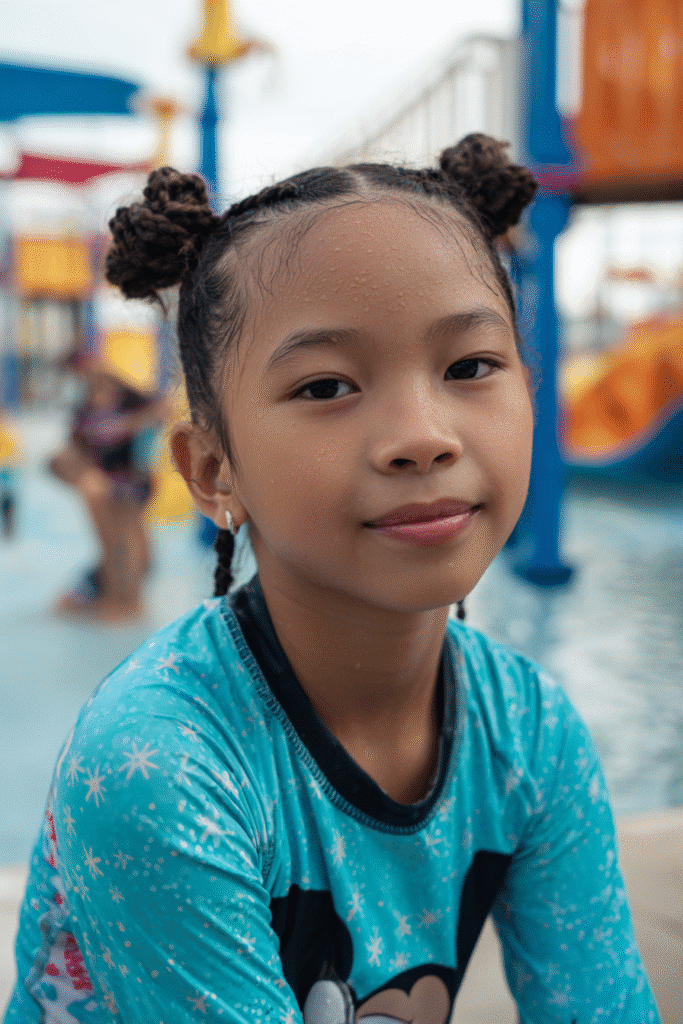 Playful American teen girl in a blue swim shirt with braided space buns, smiling in front of a splash pad at the waterpark.