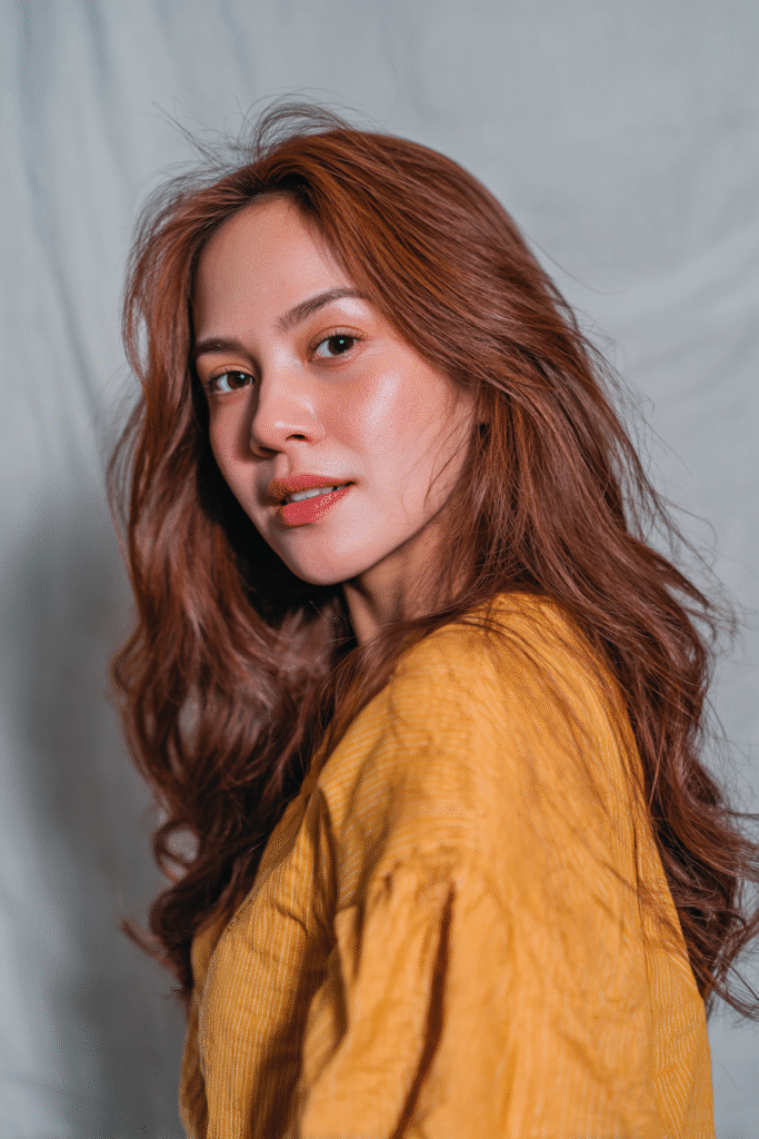 A vibrant American female model in her 20s with warm cinnamon pumpkin spice hair color, styled elegantly, photographed against a soft gray fabric backdrop with minimal texture. She has subtle makeup, a trendy outfit, a warm skin tone, and a friendly expression, captured with soft natural lighting.